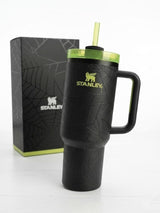 A black Stanley insulated tumbler with a handle, textured crackle pattern, and a translucent green lid with a straw, shown next to its black branded box on a white background.